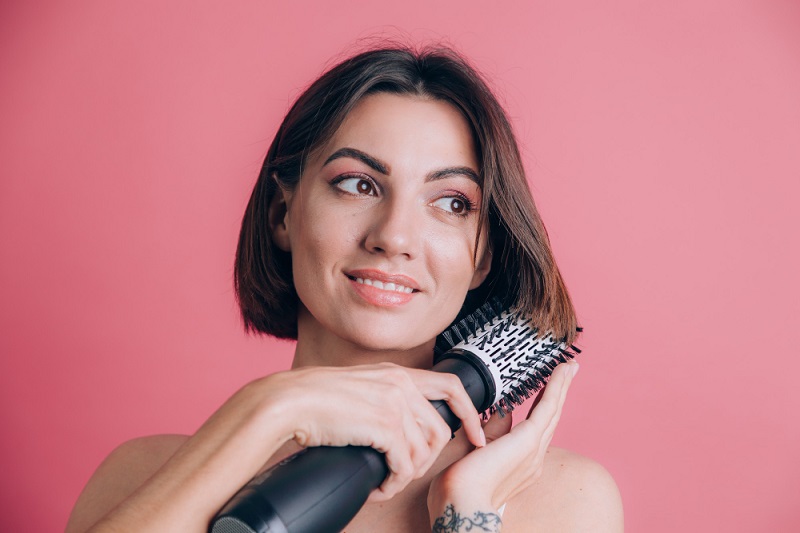 Women on pink background hold round brush hair dryer to style hair
